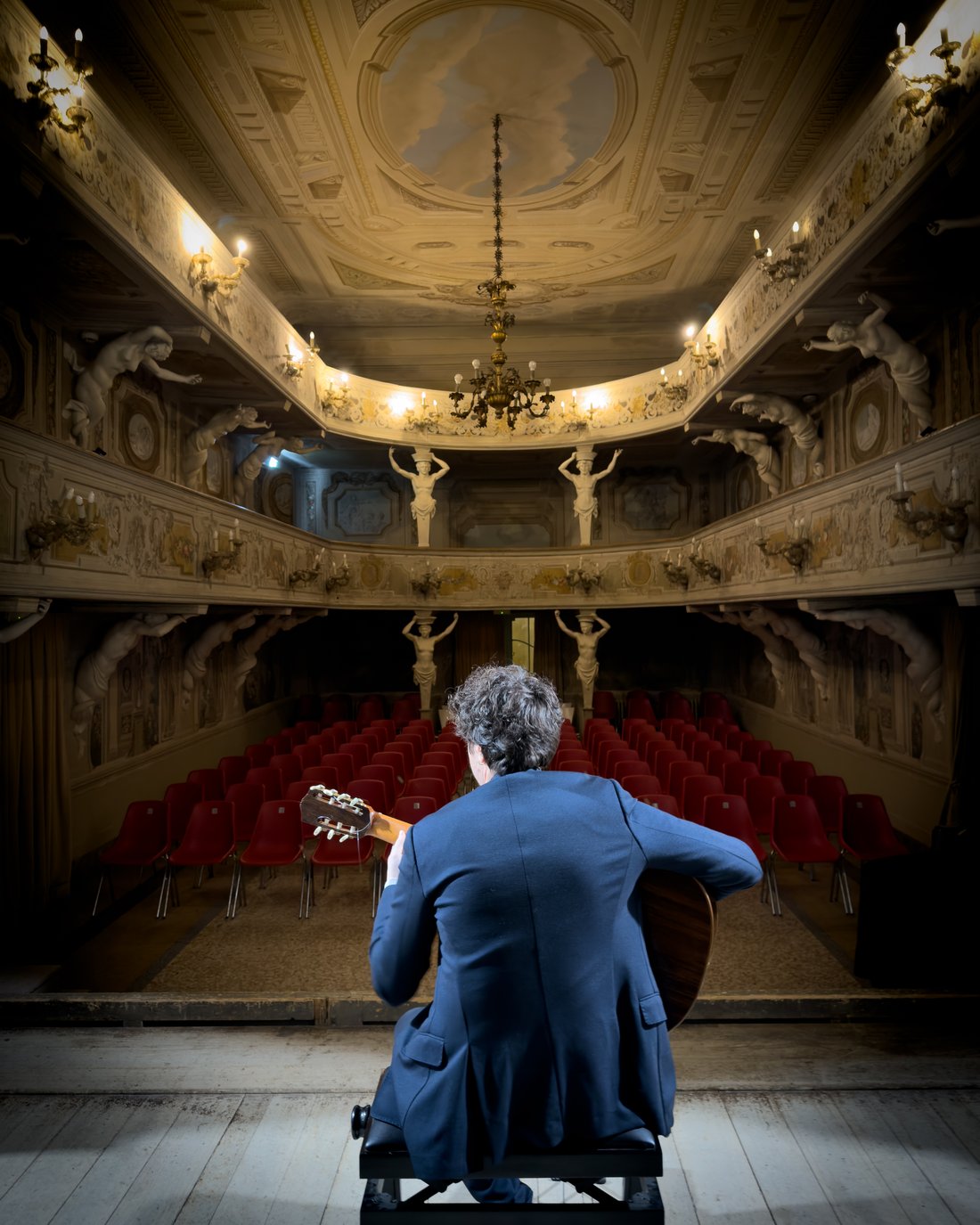 Emanuele Schillaci seated on stage with his guitar, facing the empty auditorium of a historic Italian theatre